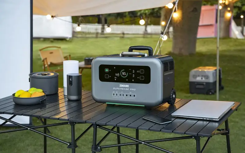 Grijs powerstation met display op een kampeertafel onder een luifel, met een laptop en lampjes